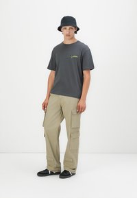 Camiseta gris con un pequeño gráfico verde, pantalones cargo beige con múltiples bolsillos y un sombrero de cubo negro. Zapatillas negras completan el atuendo.