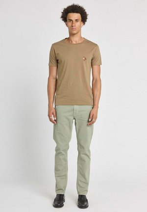 Homme aux cheveux bouclés portant un t-shirt marron avec un petit emblème de tigre et un pantalon vert clair, debout devant un fond blanc uni.