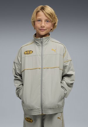 Light gray sports jacket with yellow accents and zipper, featuring Puma logo and emblem on the chest. Model stands with hands in pockets.