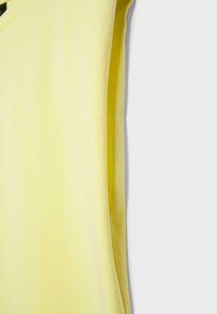 Nahaufnahme eines hellgelben ärmellosen Oberstoffs mit sichtbarer Struktur vor einem einfarbigen hellgrauen Hintergrund.