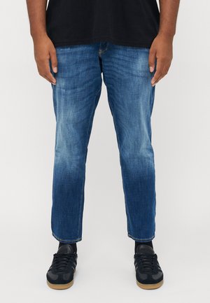Man in blauwe vervaagde rechtlopende jeans, zwarte sneakers met rubberen zolen, zwarte sokken en een zwart shirt, staande tegen een effen achtergrond.