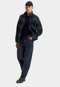 Groen fleece jack met een hoge kraag, zwart trui eronder, donkerblauwe spijkerbroek en bruine schoenen. Model staat in een ontspannen houding.