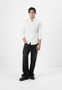 Camisa blanca de botones con mangas largas, pantalones vaqueros negros y zapatos negros. La camisa tiene un pequeño logo en el pecho. El modelo está de pie frente a un fondo blanco.
