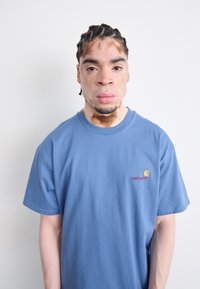 Blauwe katoenen t-shirt met korte mouwen, een ronde halslijn en een klein, geborduurd Carhartt-logo in rood en geel op de borst.