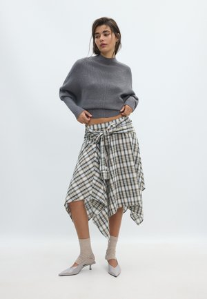 Femme portant un pull court gris, une jupe asymétrique à carreaux nouée à la taille, des chaussettes beiges et des chaussures grises pointues à petit talon, debout sur fond blanc.