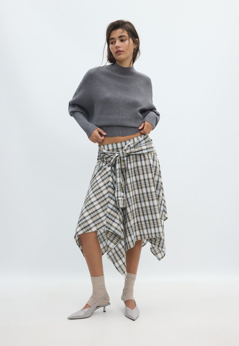 Femme portant un pull court gris, une jupe asymétrique à carreaux nouée à la taille, des chaussettes beiges et des chaussures grises pointues à petit talon, debout sur fond blanc.