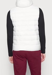 Man som bär en vit vadderad ärmlös jacka, svart långärmad skjorta och vinröda byxor, sedd bakifrån mot en enfärgad bakgrund.