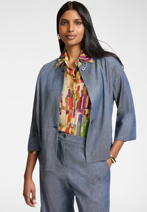 Vrouw met lang zwart haar, gekleed in een blauwgrijs tweedelig pak over een kleurrijk gestreept blouse, poseert met één hand in haar zak tegen een witte achtergrond.