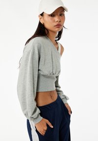 Grijze cropped trui met een enkel-schouderdesign en geplooide textuur, in combinatie met blauwe joggers met zijzakken. Witte pet als accent.