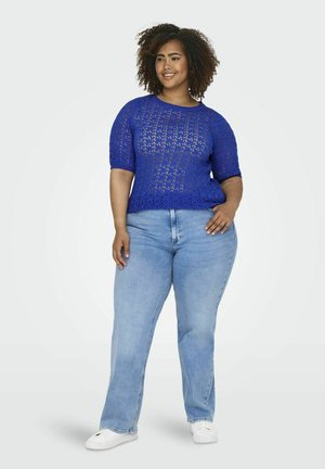 Blauwe gehaakte top met korte mouwen en een patroonstructuur, gecombineerd met lichtblauwe rechte spijkerbroeken en witte sneakers.