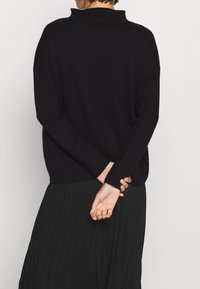 Schwarzer, lässig geschnittener Pullover mit hohem Kragen, strukturiertem Strickstoff und langen Ärmeln, kombiniert mit einem schwarzen, plissierten Rock.