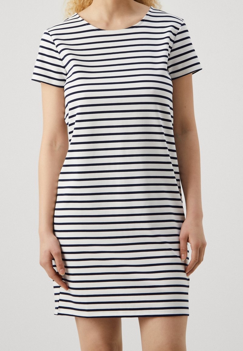 Femme portant une robe blanche à manches courtes avec des rayures horizontales bleu marine, debout devant un fond clair uni.