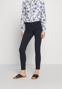 Mujer vestida con pantalones negros ajustados, una camisa blanca de manga larga con un patrón floral azul, y sandalias planas negras de dedo al descubierto, de pie frente a un fondo blanco.