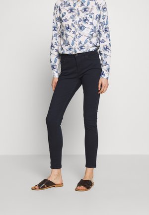 Donna che indossa pantaloni neri slim, una camicia bianca a maniche lunghe con un motivo floreale blu e sandali neri aperti nella parte anteriore, in piedi davanti a uno sfondo bianco.