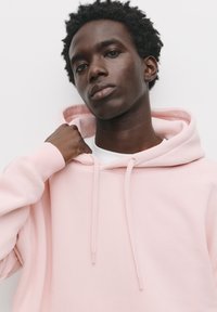 Helles rosa Hoodie mit weißem Innenfutter, Zugbändern und gerippten Bündchen. Das Modell zieht sanft die Kapuze in Richtung ihrer Schulter.