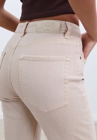 Beige denimjeans med en slät yta, med två bakfickor och en rektangulär läderlapp märkt "D98" på midjebandet.