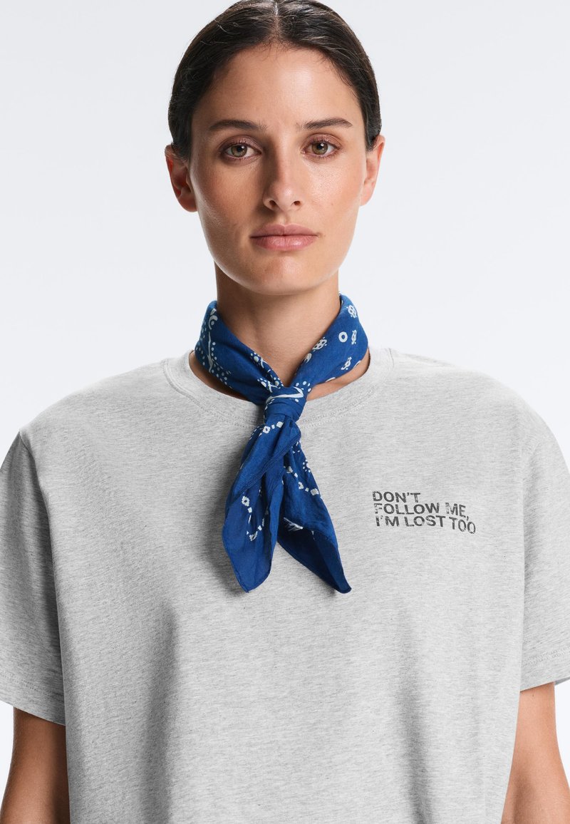 Femme aux cheveux foncés attachés, portant un t-shirt gris avec le texte « NE ME SUIVEZ PAS, JE SUIS PERDUE AUSSI » et un foulard bleu à motifs noué autour du cou.
