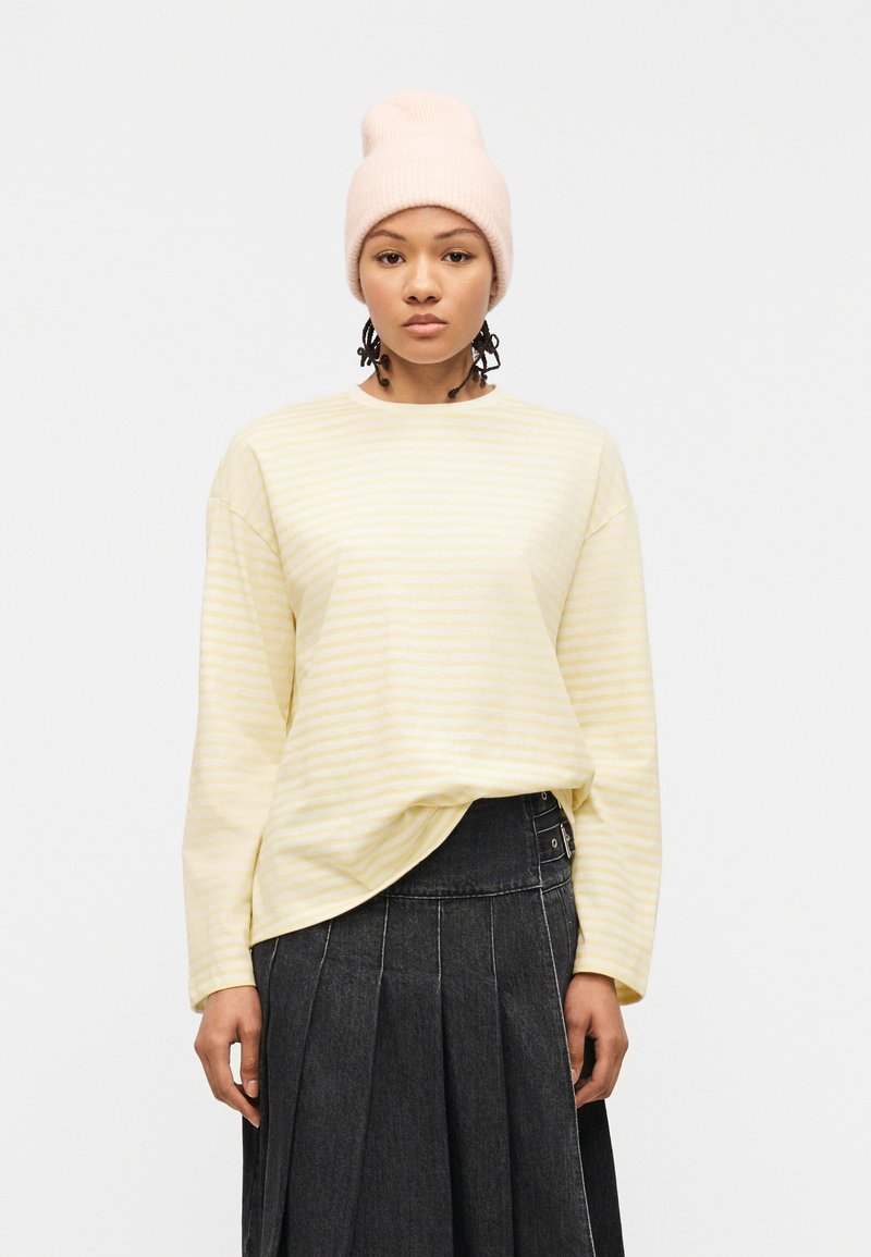 Γυναίκα φορώντας μια ανοιχτόκκινη λεπτομής ριγέ μακρυμάνικη μπλούζα, μαύρη πλισέ φούστα και ανοιχτό ροζ πλεκτό σκουφάκι, στέκεται μπροστά σε απλό φόντο.