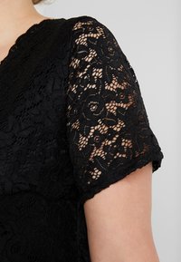 Svart spetsblus med korta, lätt puffiga ärmar. Har intrikata blommönster och en mjuk textur som framhäver genomskinliga element.