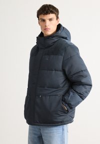 Ναυτικό μπουφάν με αποσπώμενη κουκούλα, ριγωτές λεπτομέρειες στους ώμους, τσέπες με φερμουάρ και ελαστικές μανσέτες. Διαθέτει λογότυπο στο στήθος.