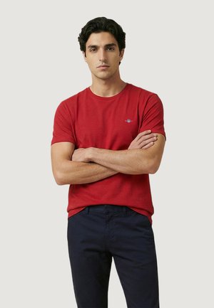 Uomo con capelli scuri, che indossa una maglietta rossa a maniche corte e pantaloni blu navy, in piedi con le braccia incrociate davanti a uno sfondo chiaro e uniforme.