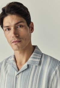 Chemise rayée en bleu clair et beige, avec un col et une fermeture par boutons, fabriquée en tissu texturé avec des détails de couture visibles.