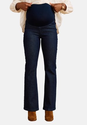 Personne enceinte portant un jean de maternité bleu foncé, remontant un t-shirt beige, et des bottines en daim marron.