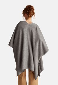 Šedý vlněný pončo s výstřihem do V a volným střihem, s texturovaným povrchem a viditelným švem, překrytý vzorovanou tkaninou pod ním.