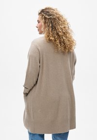 Beige neulekarte, rento istuvuus, resorireunus ja sivutaskut. Takakuvassa näkyy löysä kangas ja pehmeä tekstuuri. Pitkät hihat.