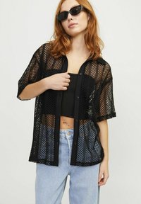 Camicia nera in rete con bottoni e maniche corte, decorata con grandi fori, indossata sopra un top aderente nero. Abbinata a jeans azzurri chiari.