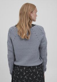 Pulover gri tricotat cu un model texturat, mâneci raglan și detalii de dantelă. Purtați peste o fustă neagră cu flori.