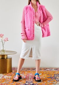 Gilet en polaire rose avec deux poches avant, associé à une jupe midi blanche à rayures avec une fente à l'avant. Sandales noires chunky avec des brides colorées.