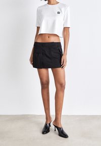 Bílý zkrácený tričko s logem na přední straně, kombinované s černou mini sukni s dvěma předními kapsami. Modelka má na sobě černé vysoké muly.