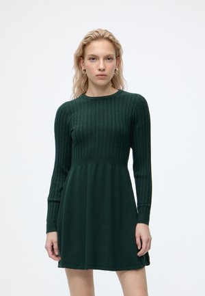 Femme aux cheveux blonds portant une robe en maille vert foncé à manches longues, debout devant un fond blanc uni.