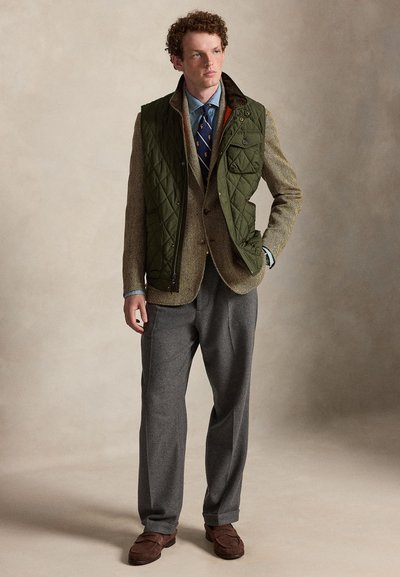 Junger Mann, der eine grüne gesteppte Weste über einem braunen Tweed-Blazer, einem blauen Hemd, gemusterten Krawatte, grauen Hosen und braunen Loafers trägt und mit der Hand in der Tasche steht.
