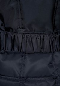 Marinblå puffjacka av tyg med en ribbad, rynkad textur vid midjan. Slät yta med sömnadsdetaljer längs sömmarna.