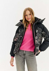 Kurtka puchowa z czarnej tkaniny z kwiatowymi wzorami w odcieniach różu i bieli, zestawiona z jasnoróżowym swetrem i szarymi jeansami.