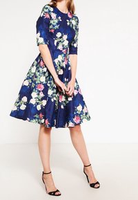 Kukka-aiheinen A-linjainen mekko tummansinisestä kankaasta, jossa on lyhyet hihat, sekoitettuja värikkäitä kukkia, kapea vyötärö ja laskostettu hame.