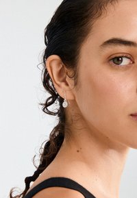 Srebrni uhani v obliki obroča s krožnim obeskom, ki ima zapletene vzorce. Model ima kodraste lase in nosi črno majico brez rokavov.