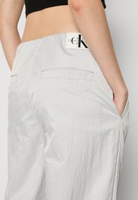 Pantalons légers gris clair, au toucher doux, avec des poches arrière et une étiquette visible de la marque Calvin Klein près de la taille.