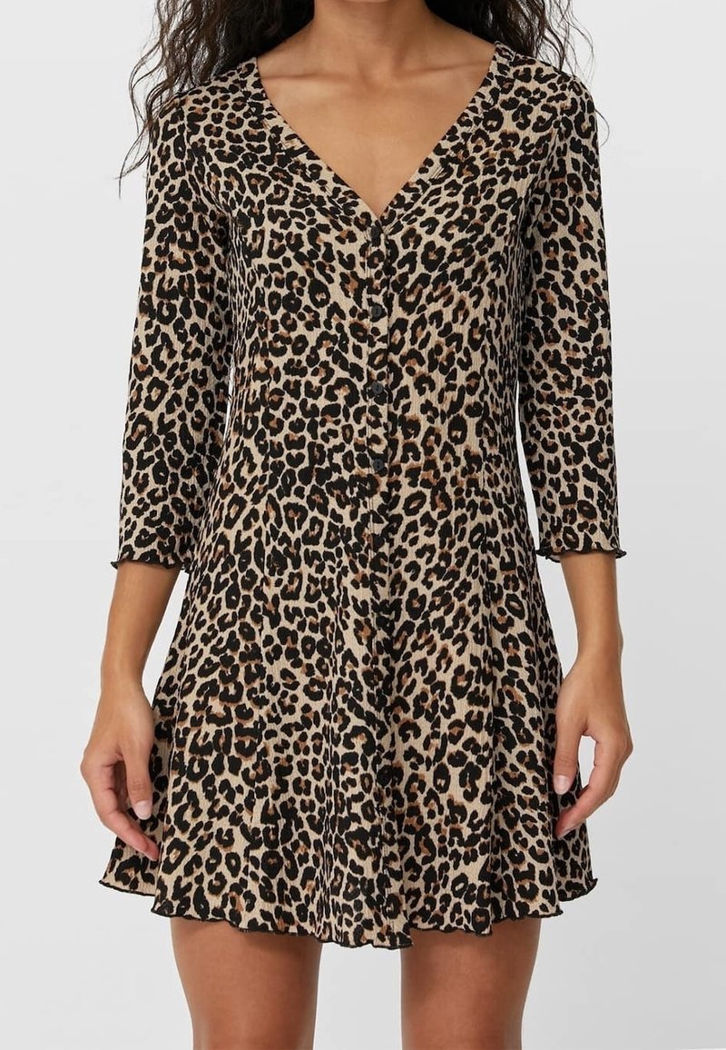 Femme portant une robe courte imprimée léopard, manches trois-quarts et col en V avec des boutons sur le devant.