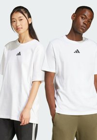 Vita bomulls T-shirts med svart Adidas-logotyp, avslappnad passform, rund halsringning, korta ärmar och enfaldig design på modellfigurer.