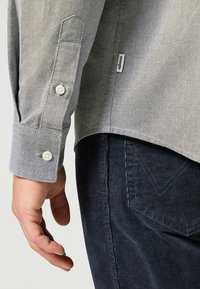 Camisa gris de botones con un acabado texturizado, puño en ángulo y botones blancos, combinada con pantalones de pana oscuros que presentan un sutil estriado.