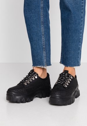Person iført sorte chunky plateau sneakers med mønstrede snørebånd og blå afkortede frynsete jeans står på en lys gulv.