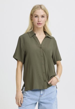 Femme blonde portant une chemise olive à manches courtes boutonnée et un jean bleu clair avec la main dans la poche sur un fond blanc uni.