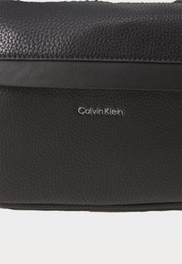 Borsa in pelle nera con una texture a ciottoli, caratterizzata da un accentuato nero liscio e dal logo argentato di Calvin Klein sul davanti. Design compatto e rettangolare.