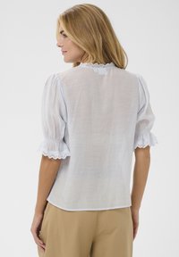Blouse blanche avec manches courtes bouffantes et bordure en dentelle festonnée, présentant un tissu léger et texturé ainsi qu'un ourlet arrondi.