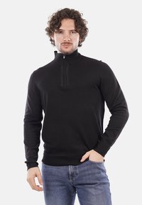 Maglione nero con zip e design testurizzato, colletto a coste. Presenta maniche lunghe e una vita aderente, abbinato a jeans blu.