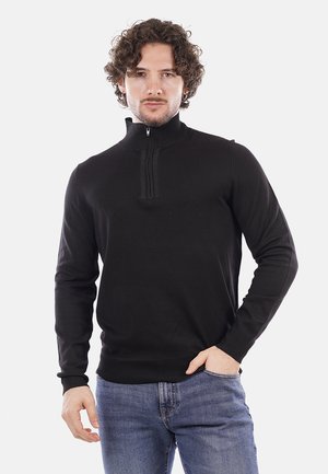 Maglione nero con zip e design testurizzato, colletto a coste. Presenta maniche lunghe e una vita aderente, abbinato a jeans blu.