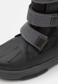 Calzado negro con una superficie texturizada, presenta amplias correas de Velcro grises y una parte superior acolchada para mayor soporte. Punta y suela reforzadas.